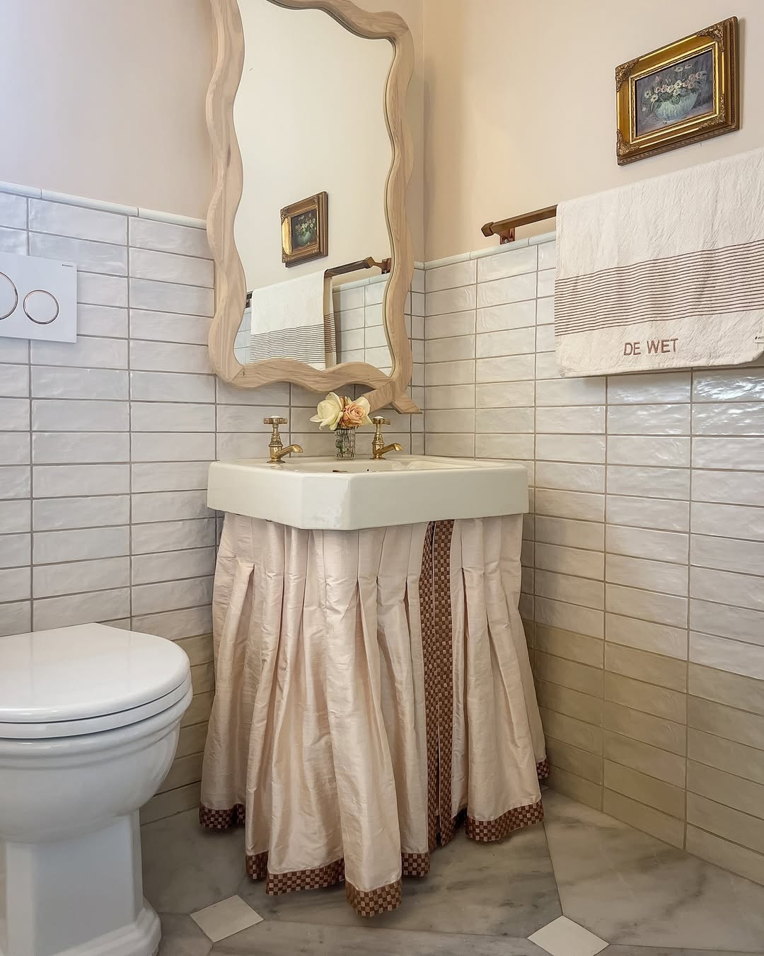 A small bathroom design featuring a white toilet, a sink with a ruffled fabric skirt, a wavy wooden mirror, a towel embroidered