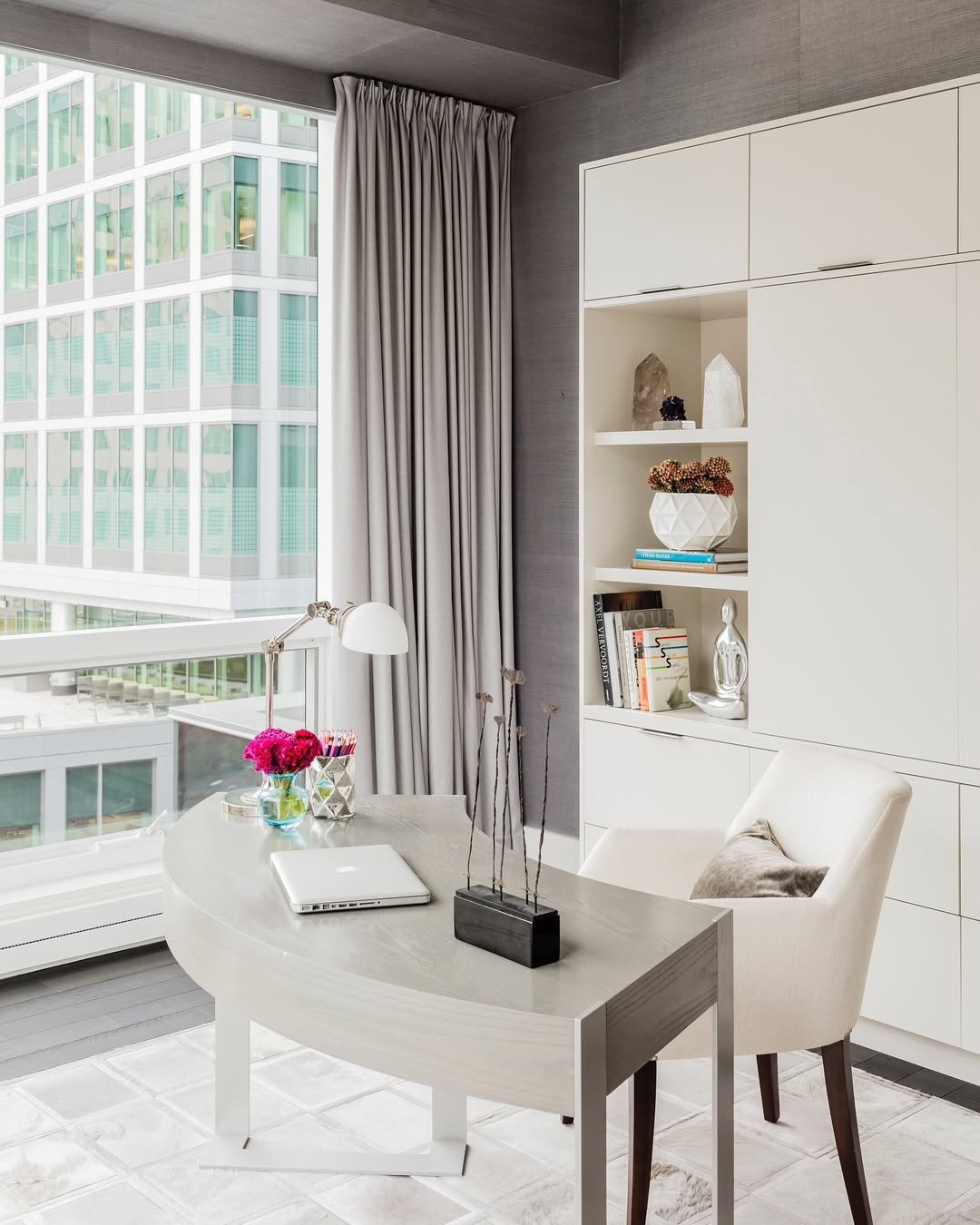 A modern small home office with a curved desk, white chair, laptop, lamp, flowers, and bookshelves. Large windows reveal an adjacent building with a grid of windows.