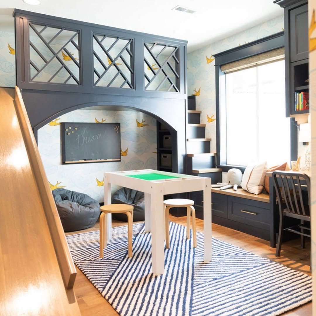 Children’s under stair playroom with a loft, slide, bean bags, a small table with stools, a chalkboard, built-in shelves, and a cozy window seat. Blue and yellow bird wallpaper decorates the walls for added charm.