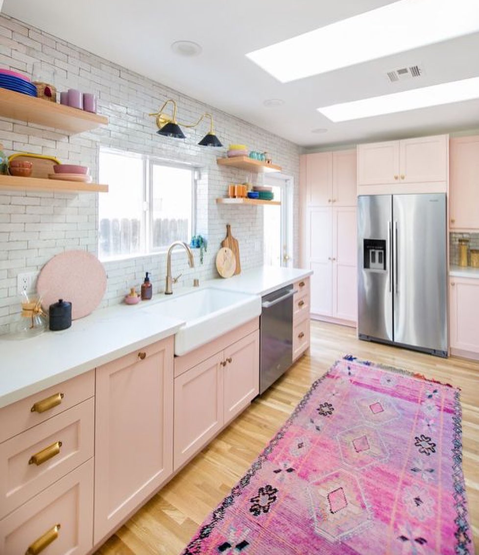 Modern pink kitchen with light pink cabinets, white countertops, stainless steel appliances, open shelves, and a pink patterned rug on a wooden floor.