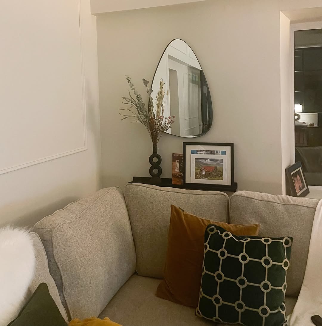 A beige sectional sofa with mustard and green pillows sits in a corner beneath a shelf holding a vase with dried flowers, framed photo, books, and an oval mirror—perfect inspiration for stylish corner decor and home decoration.