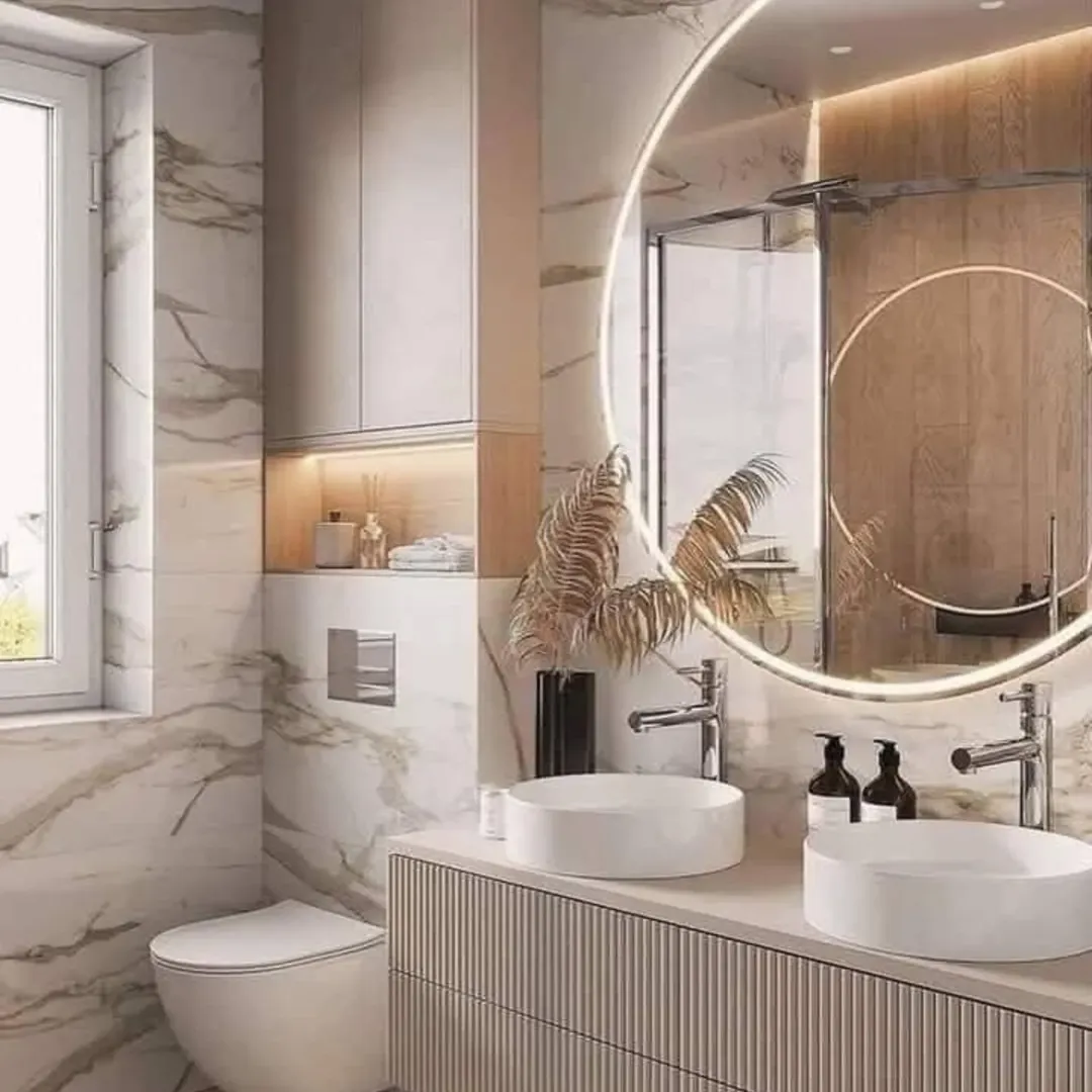 Modern bathroom design with marble walls, double sinks, round backlit mirror, glass shower, and decorative dried plants beside the faucets—perfect for those seeking small bathroom decor inspiration.