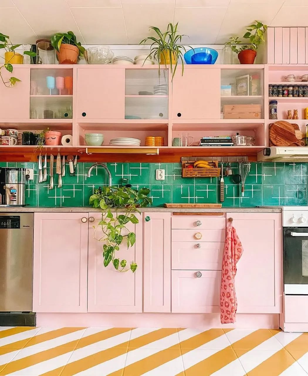 A charming pink kitchen featuring pink cabinets, a green tiled backsplash, open shelving with dishes and plants, and a vibrant diagonal yellow-and-white striped floor.