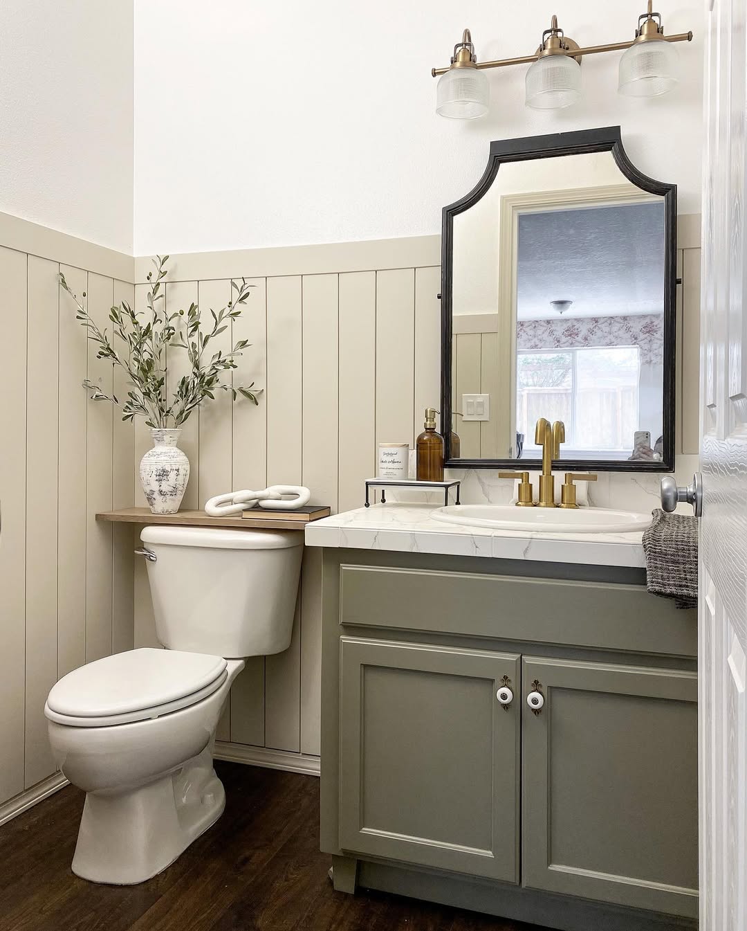 Discover inspiring half bathroom ideas in this inviting space featuring a green vanity, white sink, wall-mounted mirror, light fixture, potted plant, and wood floor. Beige panel walls with a white upper section add charm to this small bathroom decor.