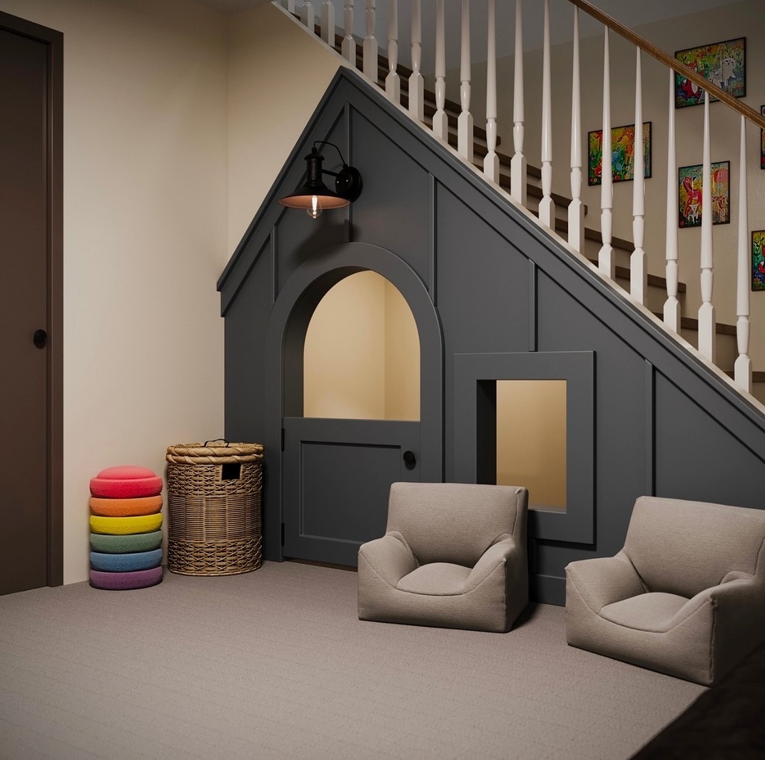 An under stair playroom features a small gray playhouse, two beige chairs, a wicker basket, stacked colorful cushions, and framed art on the walls—creating a cozy hideaway for kids.