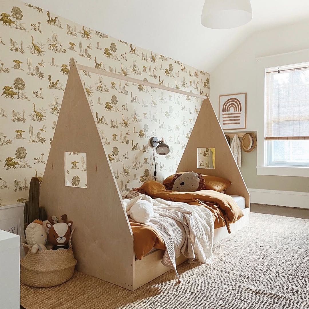 A dinosaur-themed children's bedroom features a wooden tent-shaped bed, yellow bedding, stuffed animals, and nature-inspired wallpaper. A wall-mounted lamp and framed dino art add charm as natural light floods in through a window.