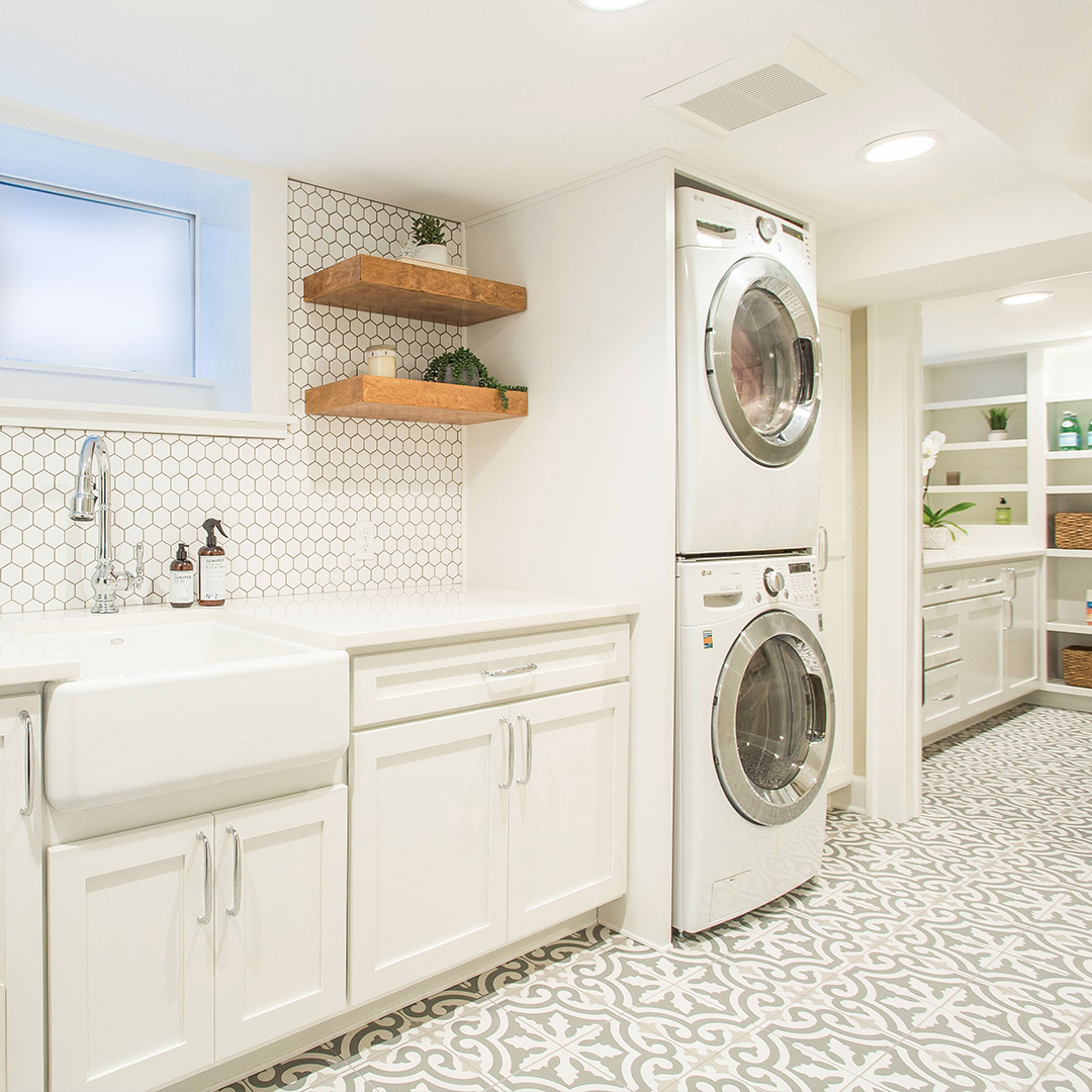 Modern cottage laundry room with white cabinetry, patterned floor tiles, a farmhouse sink, stacked washer and dryer, open wooden shelves, and decorative plants.