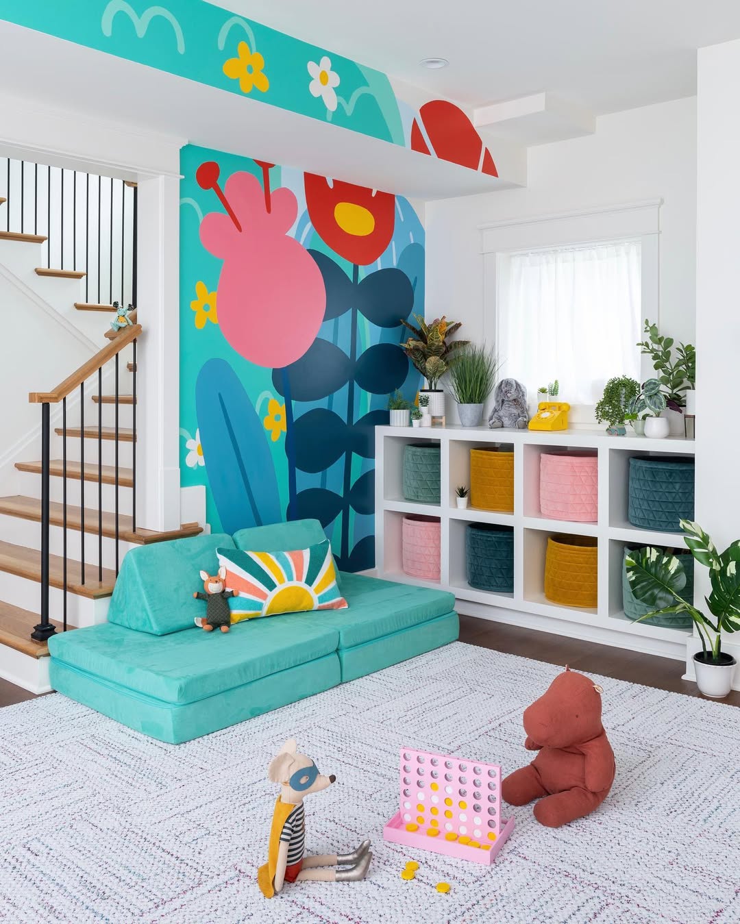 A brightly decorated under stair playroom with a colorful mural, a teal couch, stuffed animals, pastel storage baskets, and a white shelf filled with plants and toys.