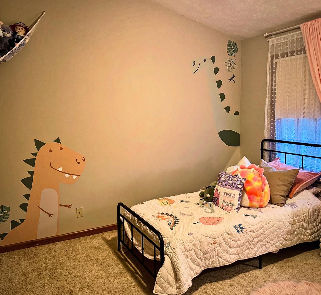 A child's dinosaur-themed bedroom with vibrant wall decals. The bed, adorned with colorful pillows and an animal quilt, is perfectly set against the wall. To the right, sheer curtains gently frame the window.