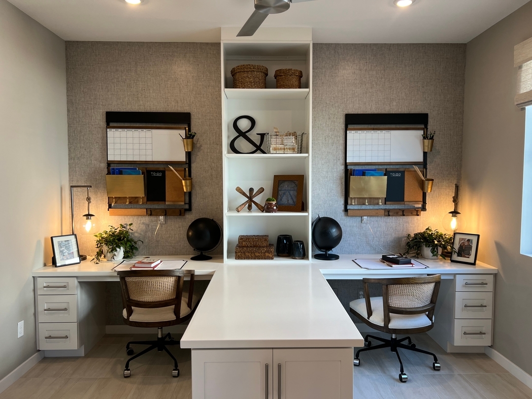 A modern small home office with two workstations, wicker chairs, wall organizers, plants, and shelves displaying decorative items, separated by a central island.