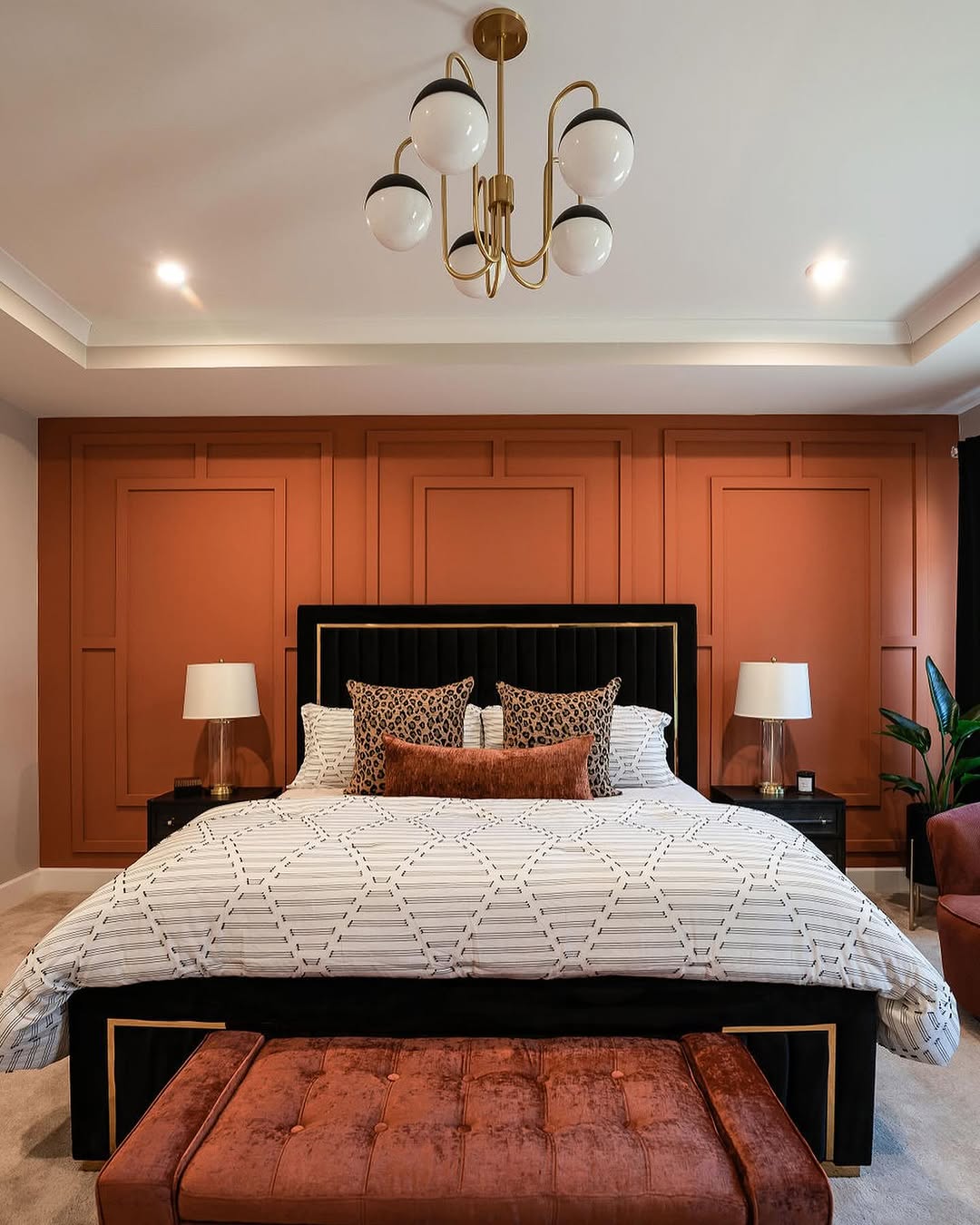 A modern bedroom with a large bed, patterned pillows, two bedside tables with lamps, a brown bedroom accent wall, a tufted bench, and a gold ceiling light fixture.