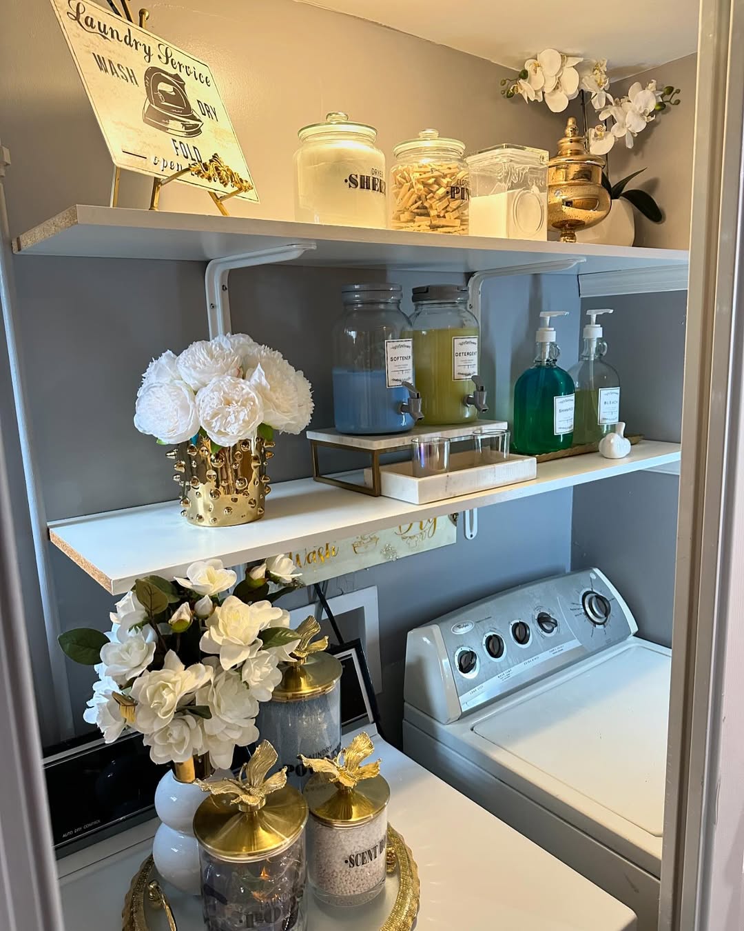 A neatly organized laundry room with a top-load washer and stylish Laundry Room Shelves, white and holding detergent, cleaners, jars, and decorative flowers—all beautifully arranged for a tidy, visually appealing space.