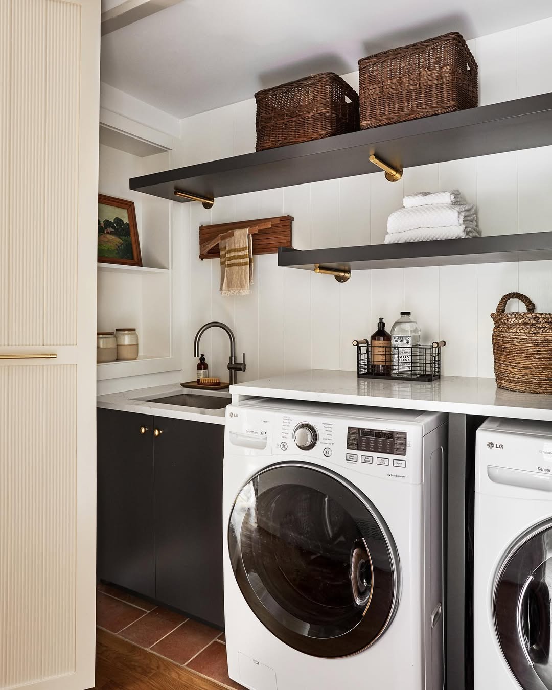 A modern laundry room with a front-loading washer and dryer, a countertop with a sink, and stylish laundry room shelves holding baskets, towels, and decor items.