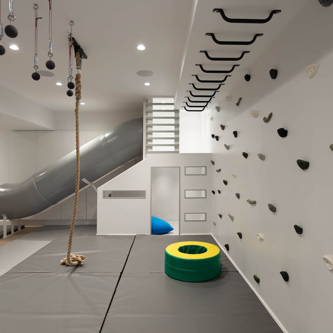Indoor under stair playroom with climbing holds on the wall, monkey bars on the ceiling, a slide, rope, padded flooring, and foam mats.