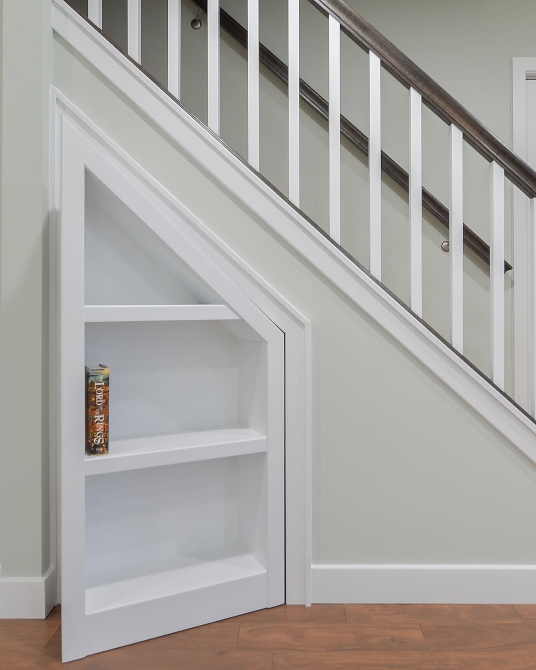 A built-in triangular white bookshelf with three shelves is fitted under a staircase, offering stylish homeowners clever hidden door ideas. Only one book rests on the bottom shelf.