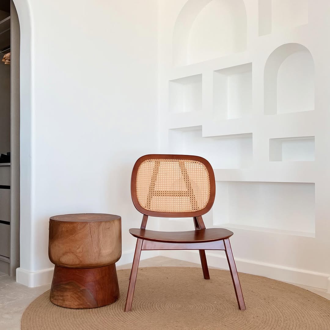 A wooden accent chair with a woven rattan backrest sits beside a round wooden side table on a circular rug—an inspiring home decor idea for minimalist interior design with white built-in wall shelves.