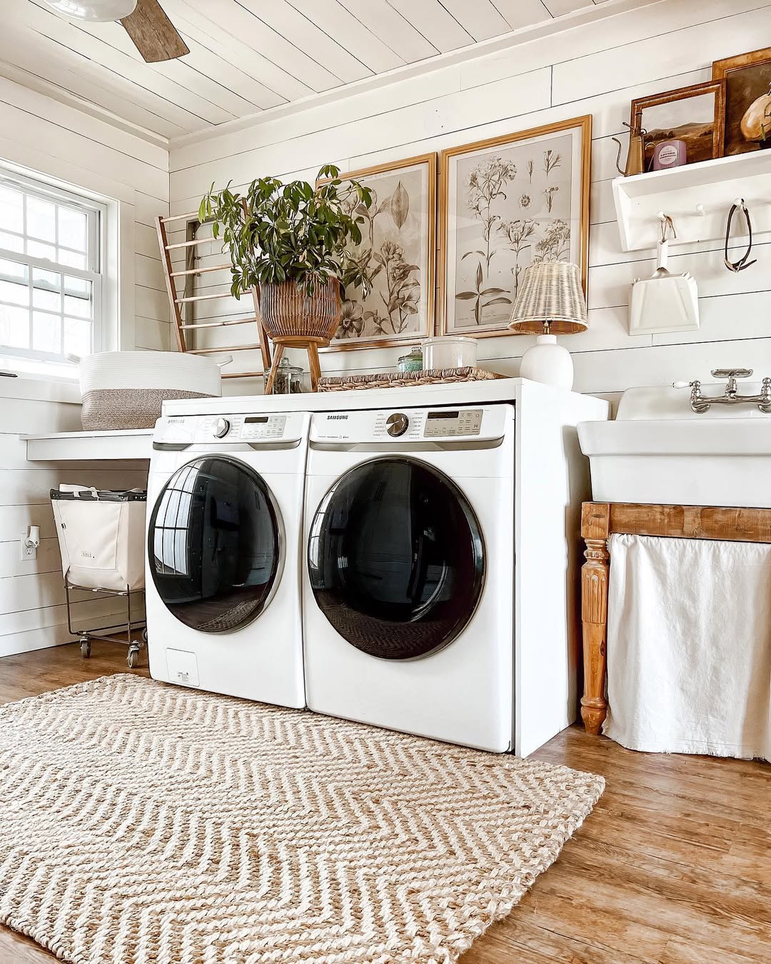 A charming cottage laundry room with a front-load washer and dryer, farmhouse sink, patterned rug, potted plant, and botanical artwork creating a cozy, inviting space.