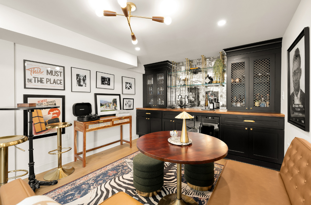 A modern home bar with black cabinetry, a round wooden table, tan chairs, wall art, zebra-patterned rug, and a gold chandelier in a well-lit room—perfect inspiration for stylish home bar designs.