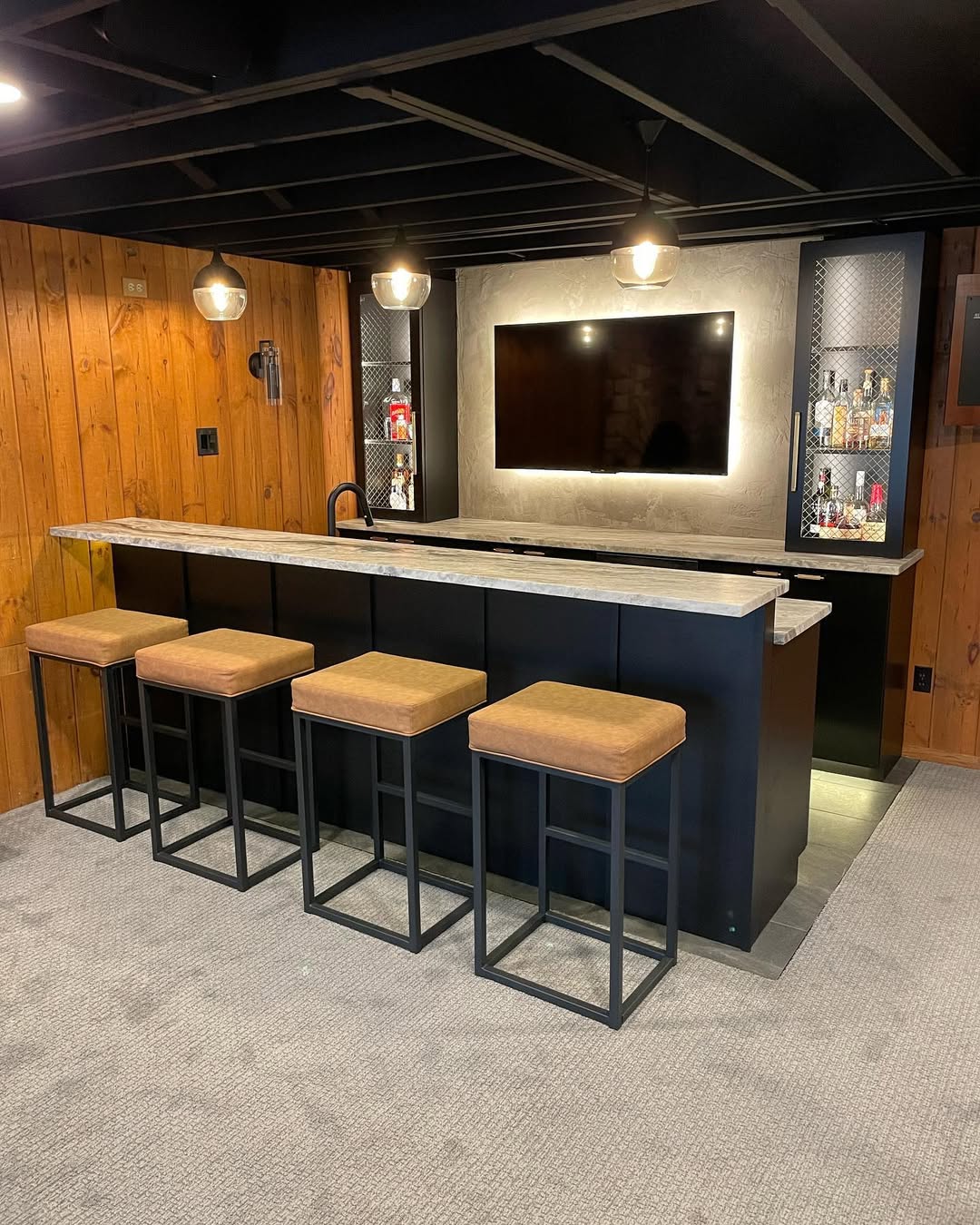 A home bar with a marble countertop, four bar stools, a wall-mounted TV, wood paneling, and shelves with bottles displayed behind the bar—one of the most gorgeous bar ideas for home entertaining.