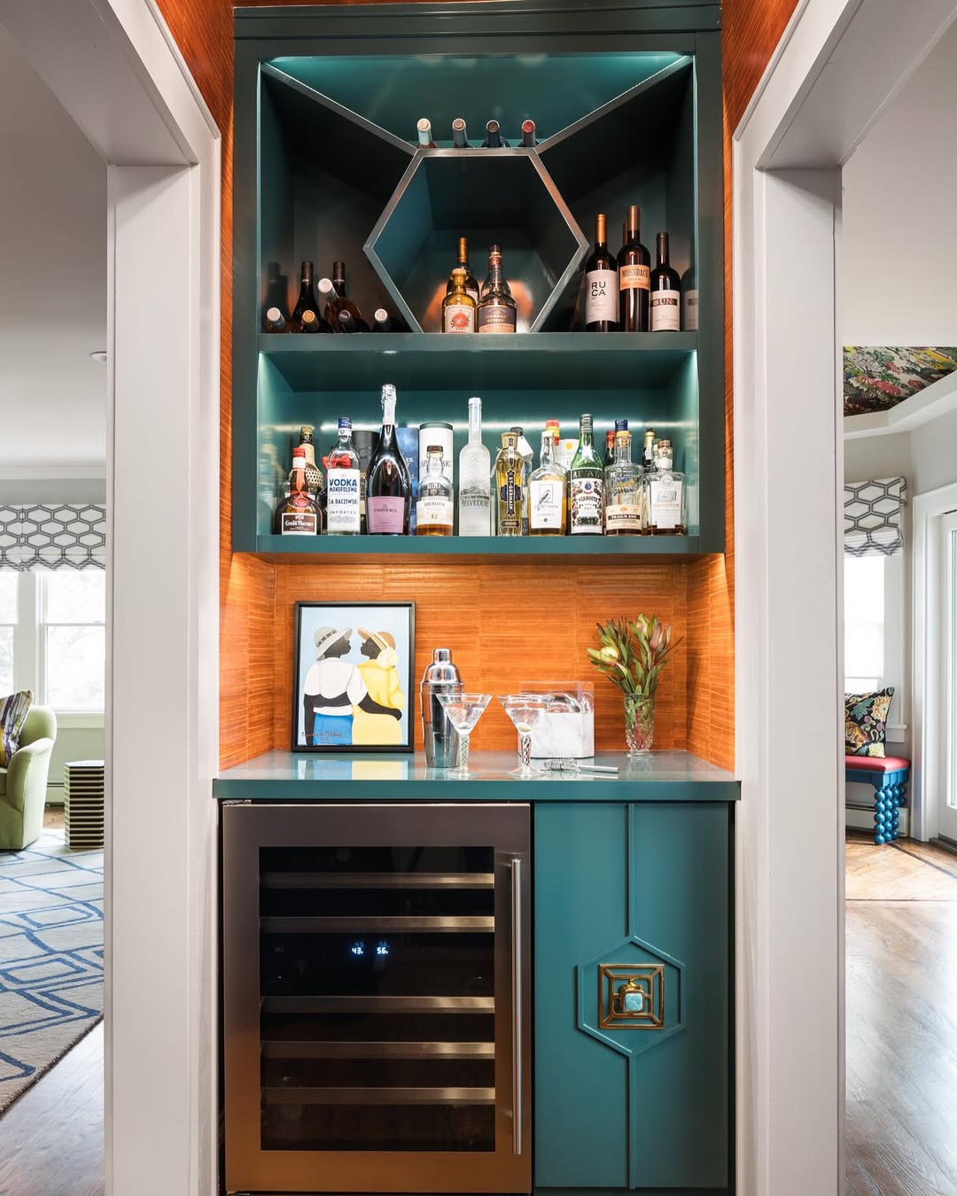 A small home bar with teal cabinets, a wine fridge, assorted liquor bottles, glasses, artwork, and a vase with flowers on the counter—perfect for those seeking gorgeous bar ideas for home.