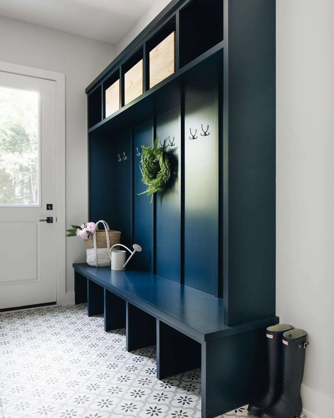 Navy blue mudroom bench with cubby storage, coat hooks, and overhead baskets offers stylish entryway organization; a potted plant, watering can, and boots sit nearby on the patterned tile floor for fresh mudroom ideas.