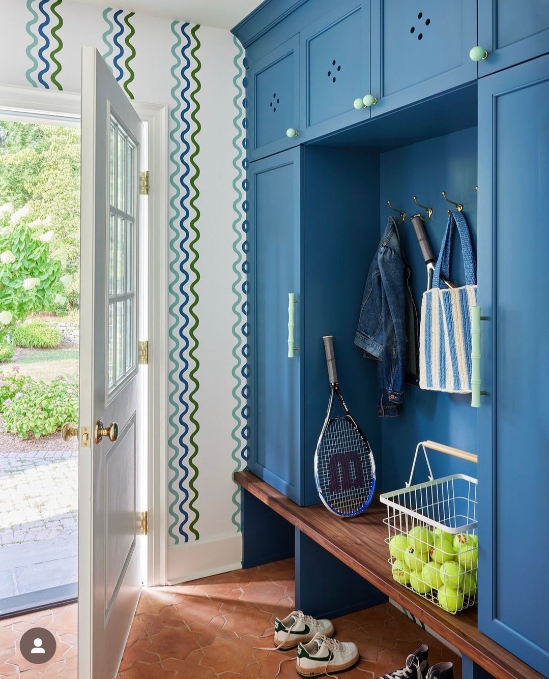 A blue mudroom with built-in cabinets, a bench, tennis gear, shoes, hanging jackets, a striped tote, and wavy wallpaper offers genius mudroom ideas near a door open to a garden.
