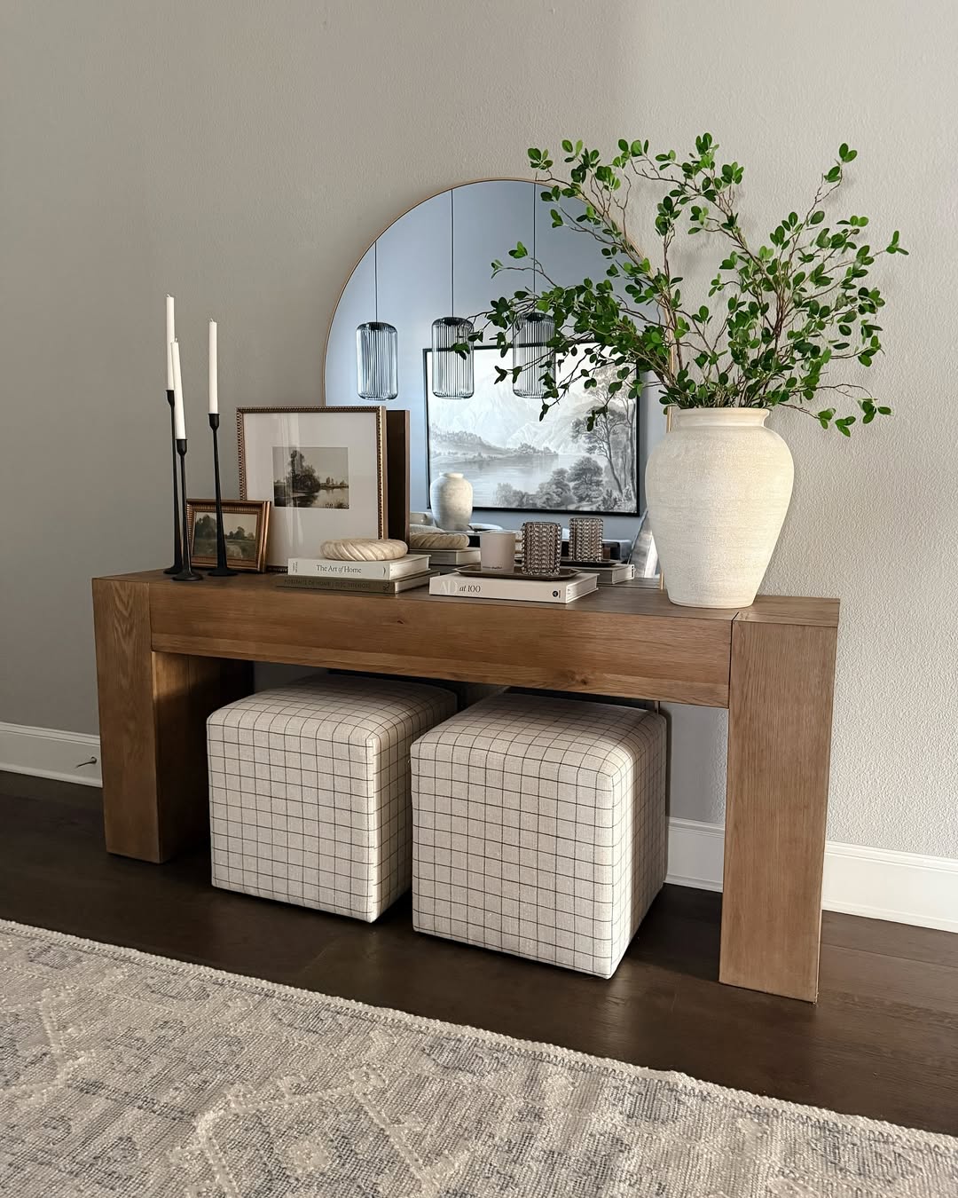 A wooden console table with a round mirror, two lamps, a vase of white flowers, candles, a woven basket, and decorative pillows underneath—perfect for inspiring stylish console tables and fresh console table decor ideas.