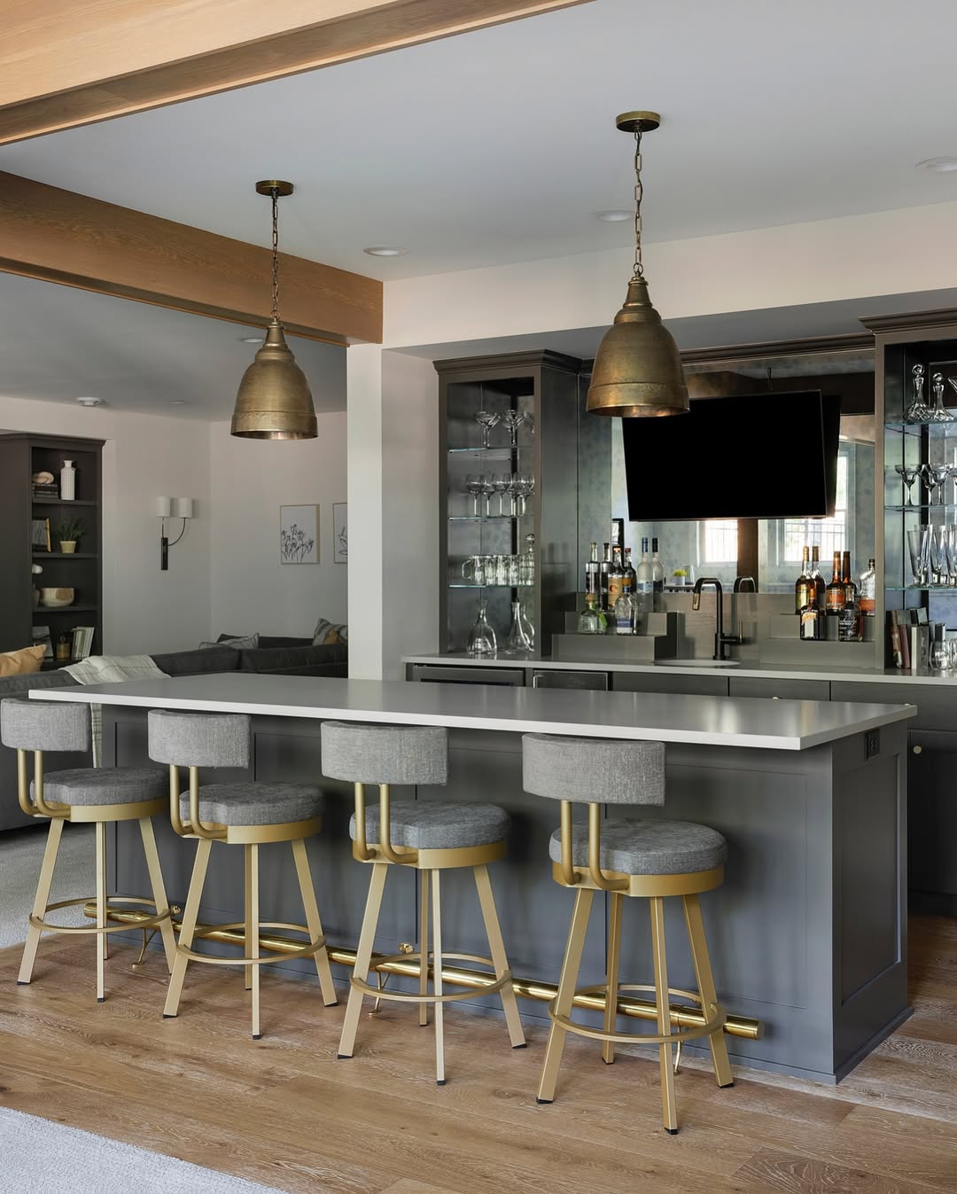 Modern home bar area with a gray island, four upholstered bar stools with gold accents, pendant lights overhead, and shelves stocked with glassware and bottles—perfect inspiration for those seeking stunning home bar ideas.