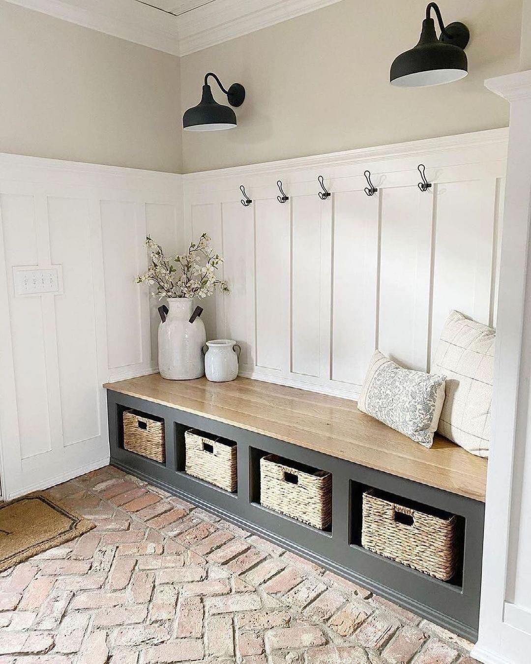 A genius mudroom design with a built-in bench, four storage baskets, wall hooks, a vase with flowers, pillows, and two black wall lamps—perfect entryway inspiration with brick flooring and white panel walls.