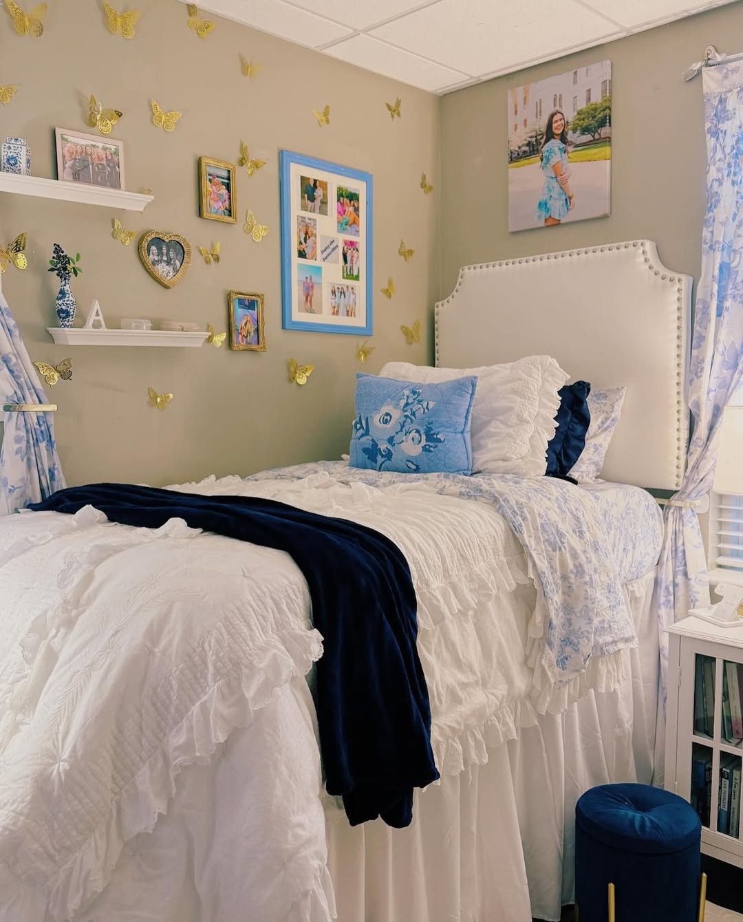 A neatly made bed with white and blue bedding, butterfly wall decorations, framed photos, shelves with decor, and a window with floral curtains create a cozy dorm room for college guys.