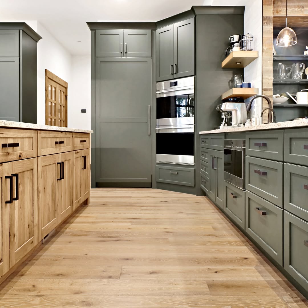 Modern kitchen with green cabinetry, wood-paneled island and floor, stainless steel appliances, open shelving, and a neutral color palette.