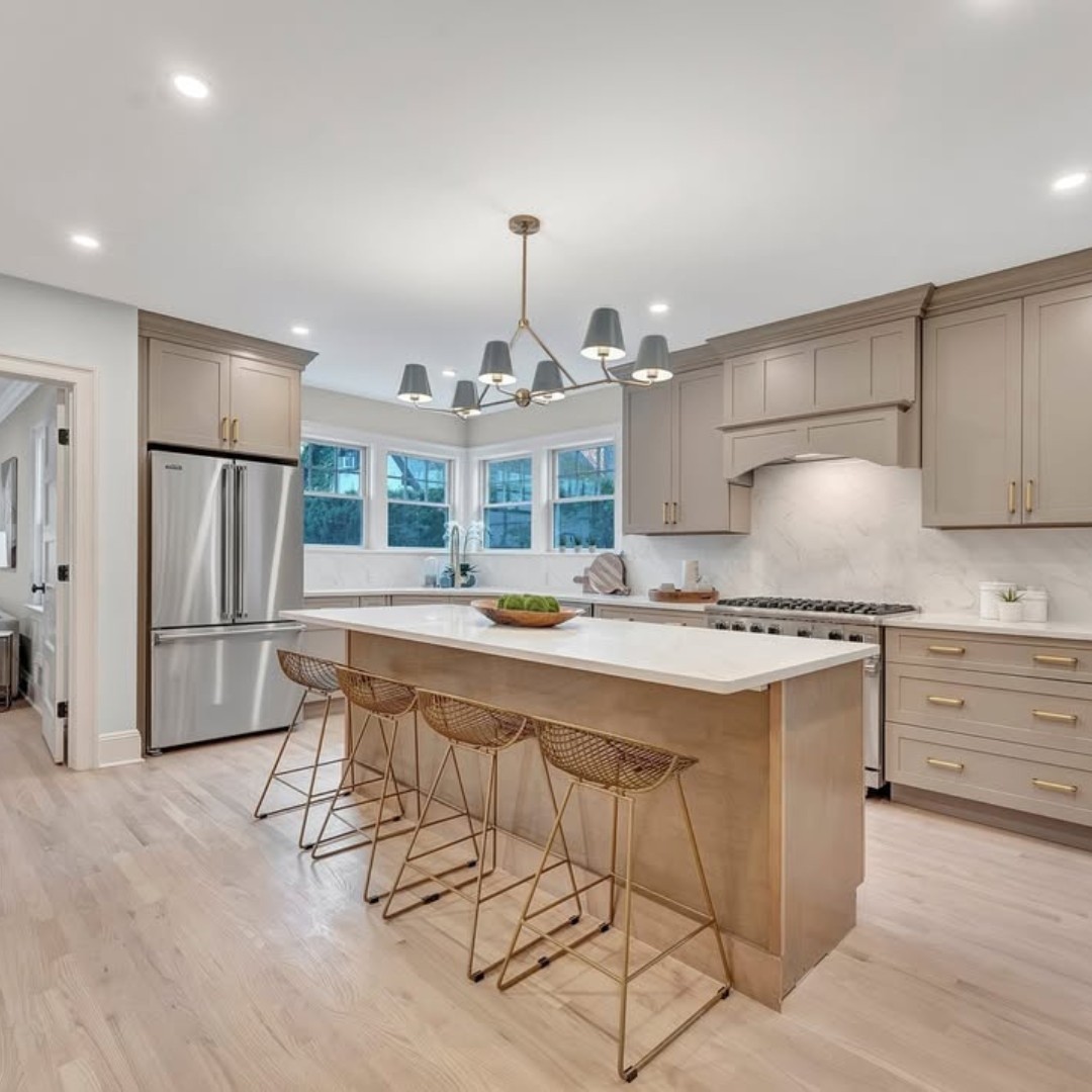 Modern kitchen with beige cabinets, stainless steel appliances, large island with marble countertop, gold accents, and three wicker bar stools under pendant lighting.