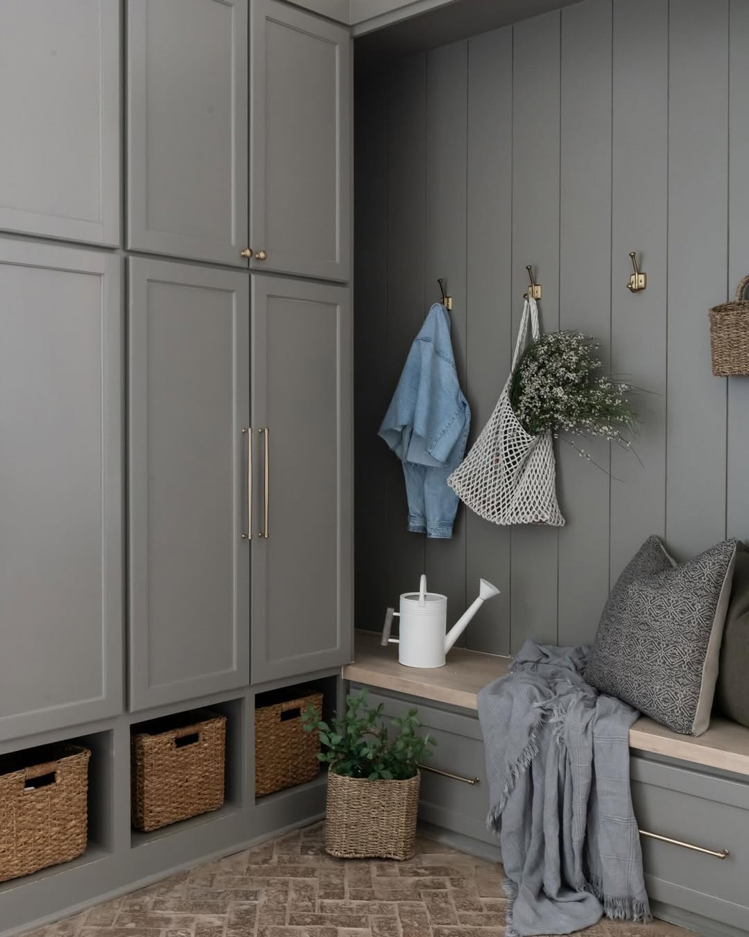 A genius mudroom with gray built-in cabinets, a bench with cushions and throw, wicker baskets, wall hooks holding a blue shirt, a mesh bag, a plant, and a white watering can creates inspiring entryway organization.
