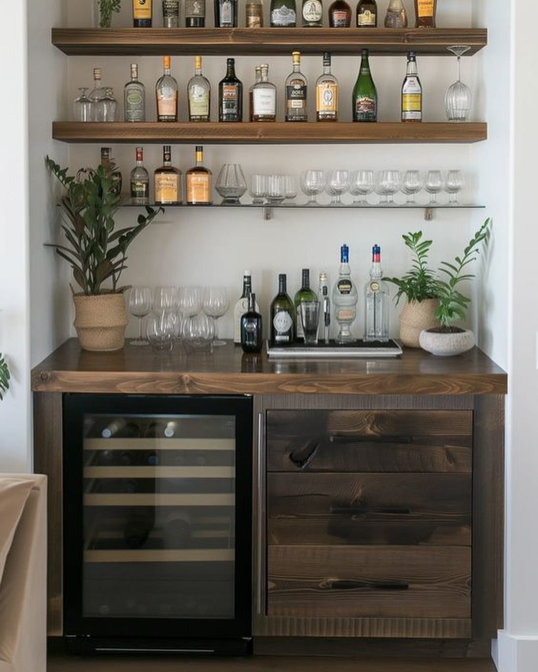A gorgeous home bar with wooden shelves holding liquor bottles and glassware, a small fridge, drawers, countertop spirits, and potted plants—a perfect source of bar ideas for home enthusiasts.
