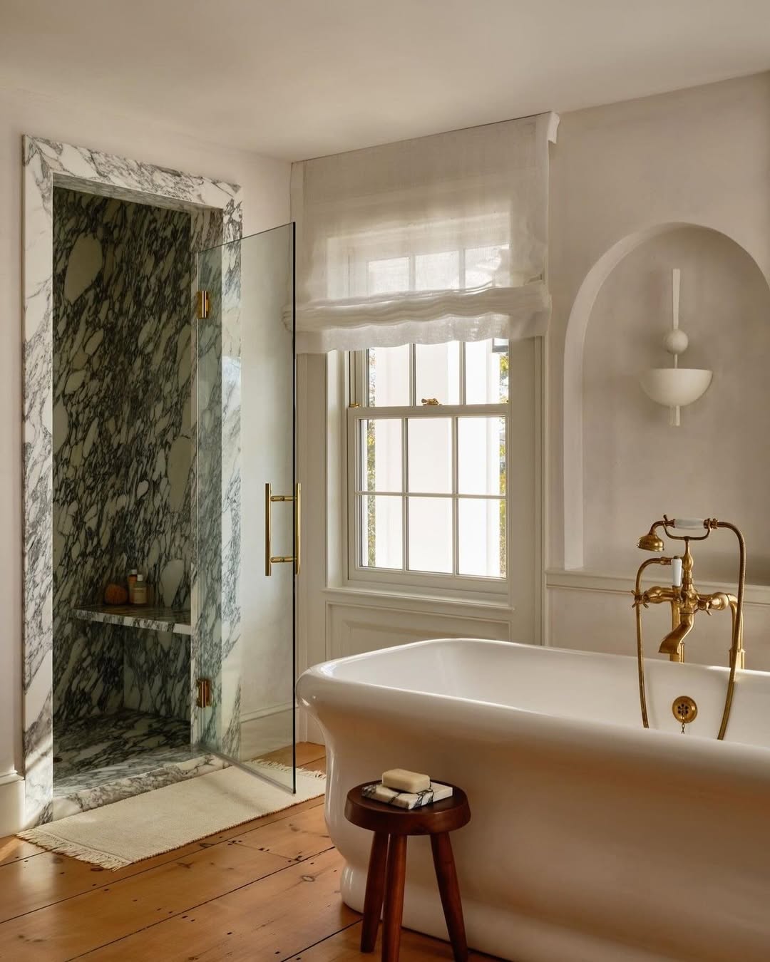 Bathroom with a freestanding white tub, brass fixtures, marble design shower, glass door, wooden stool with soap, wood floor, and a window with a sheer shade—perfect inspiration for elegant bathroom ideas.