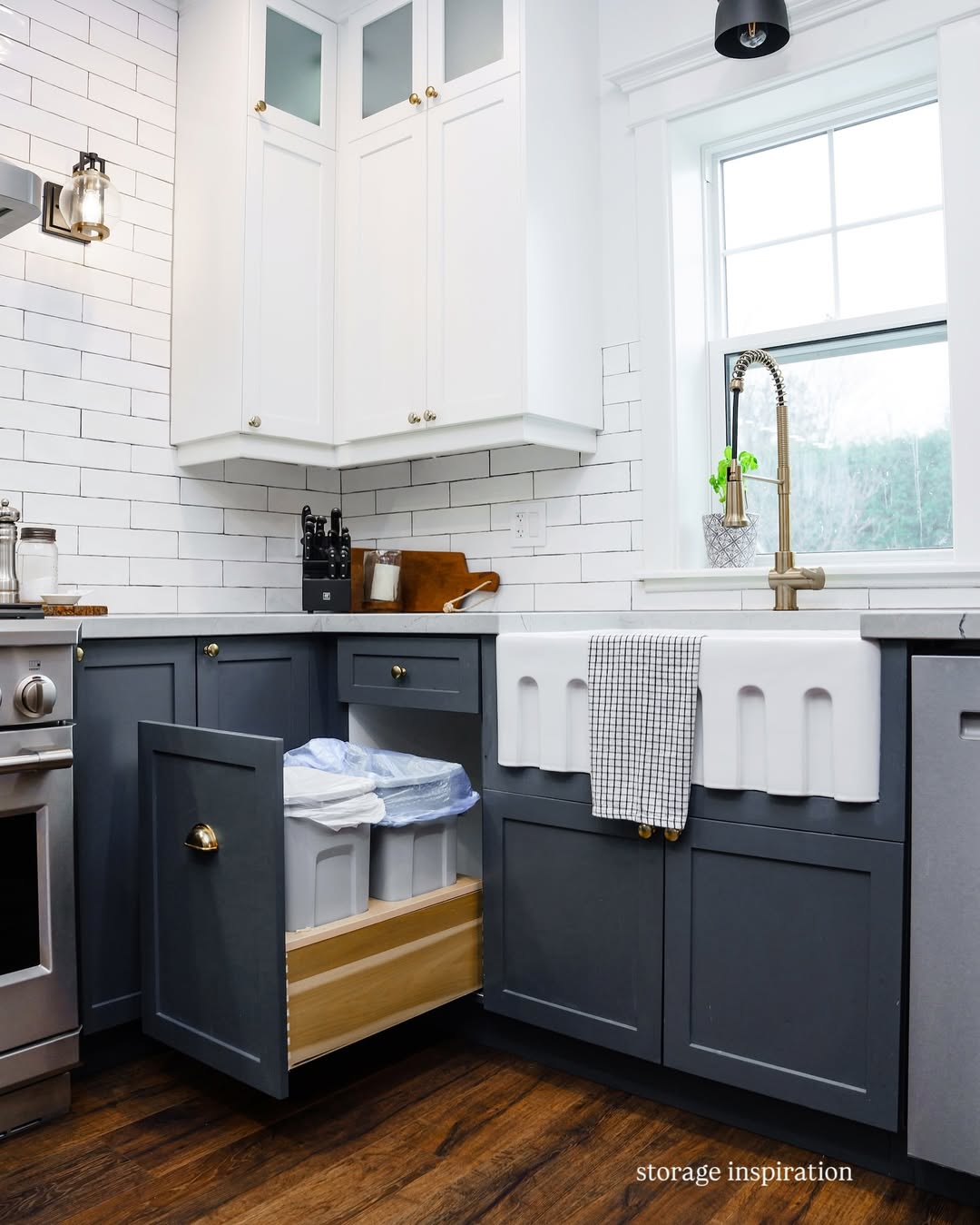Modern kitchen with white upper and dark lower cabinets, a farmhouse sink, white subway tile backsplash, and a pull-out drawer storing bins and towels under the counter.