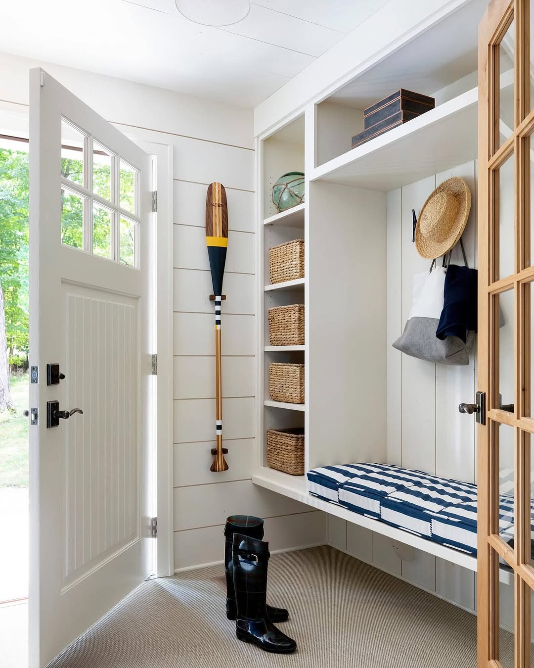 A stylish mudroom features a patterned bench, wicker baskets on shelves, a hanging hat and bag, a wooden paddle on the wall, and rain boots by the open door—perfect inspiration for functional mudroom ideas.
