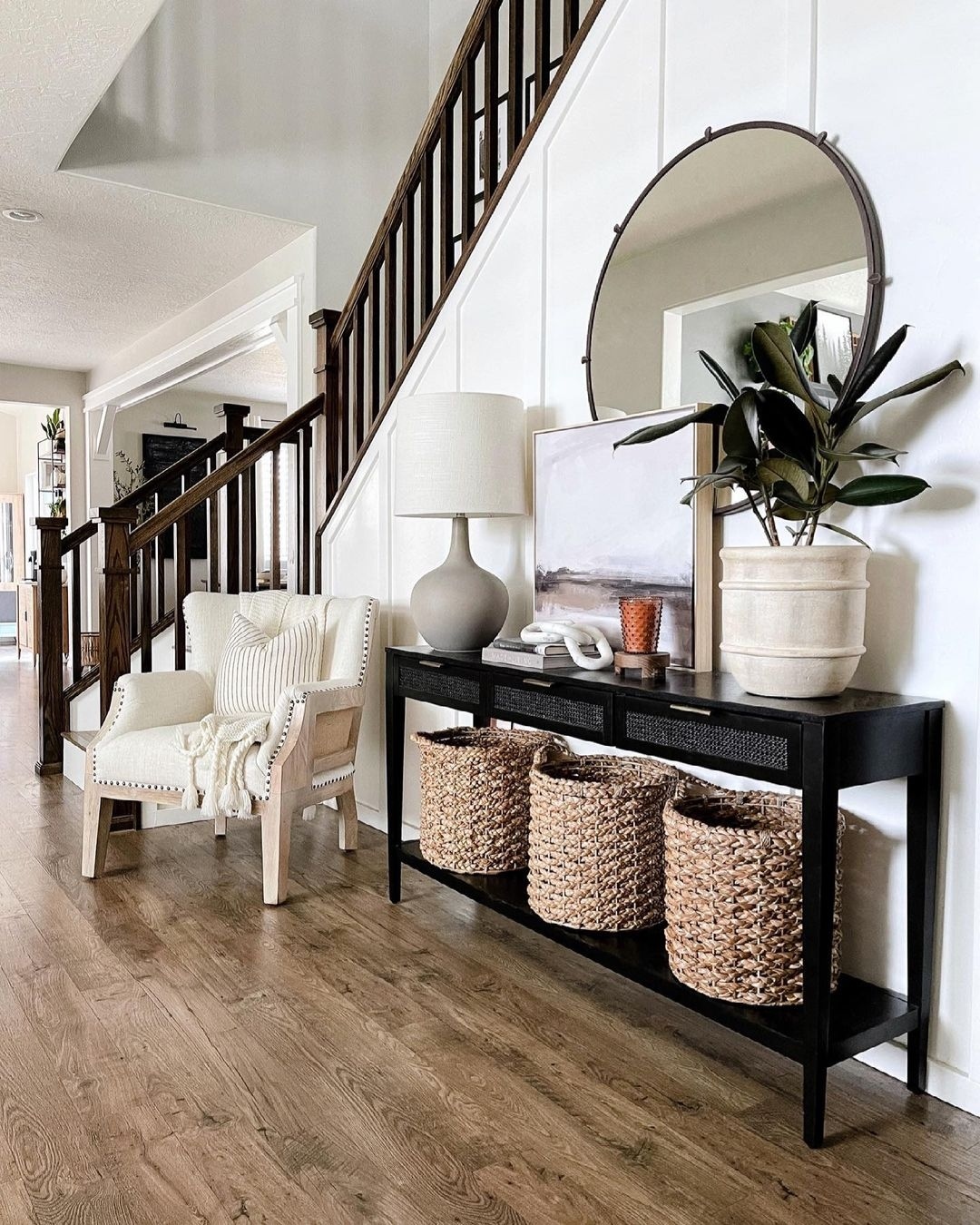 Entryway with stylish console table decor, woven baskets, potted plant, lamp, framed art, round mirror, and a cream armchair beside a stairway with dark wood railings.