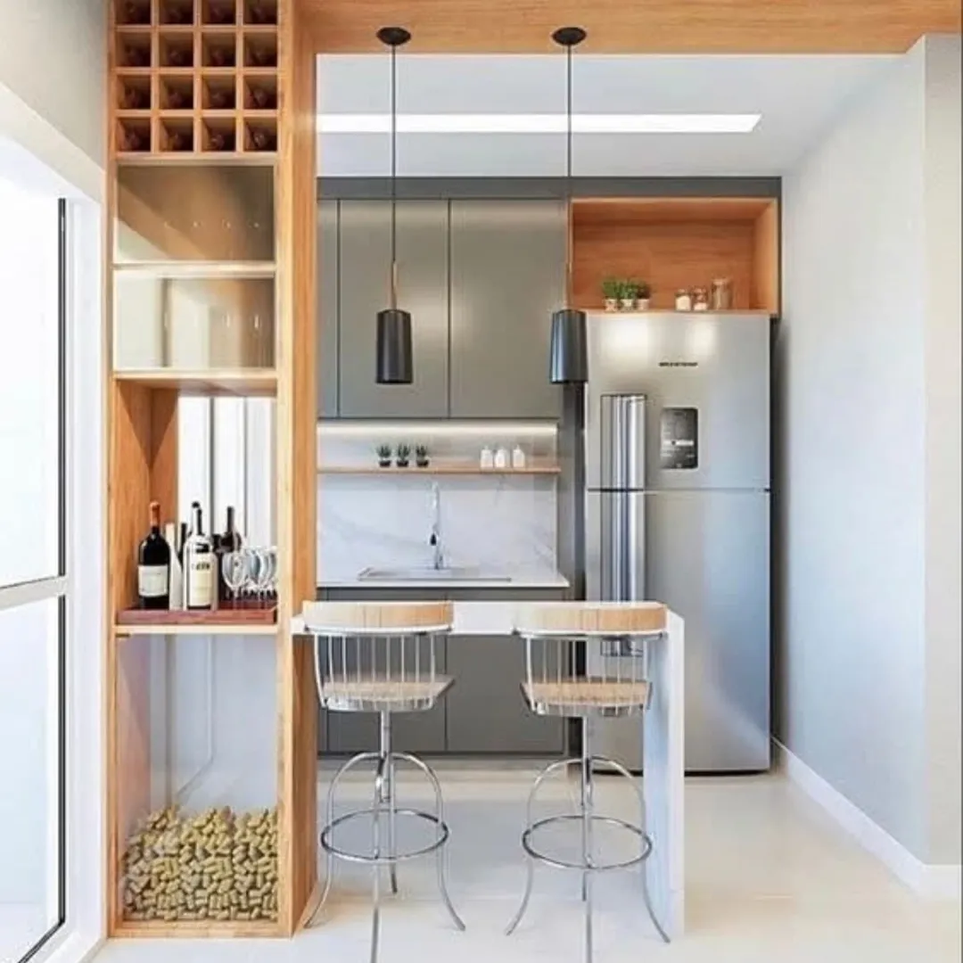 Modern kitchen with gray cabinets, a stainless steel fridge, a small breakfast bar with two chairs, pendant lights, and wooden shelves with wine storage and glassware.
