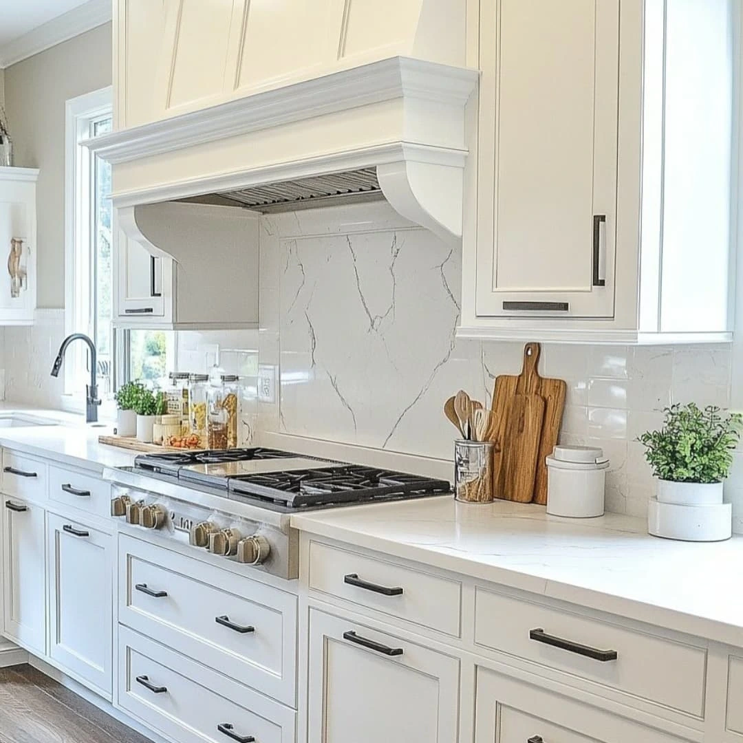 A modern white kitchen features a gas stove, marble-look backsplash, white cabinets with black handles, and countertop accessories including cutting boards, jars, and potted herbs.