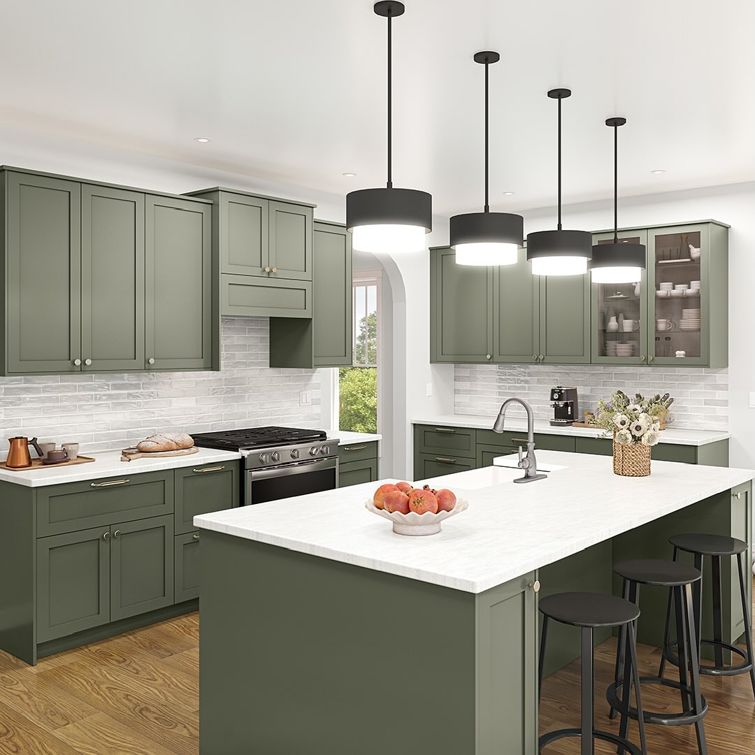 Modern kitchen with sage green cabinets, white countertops, an island with black stools, and three pendant lights above. There are flowers and a bowl of peaches on the island.
