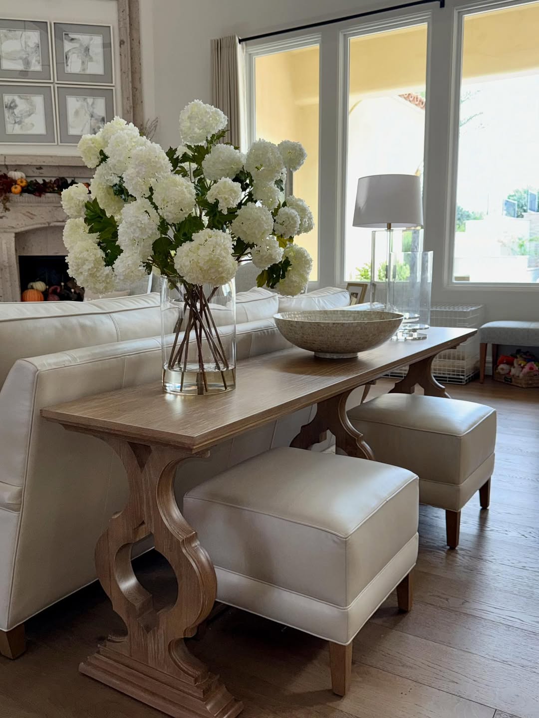 A stylish console table with a vase of white flowers, a decorative bowl, and two stools is placed behind a white sofa, offering inspiring console table decor in a well-lit living room.