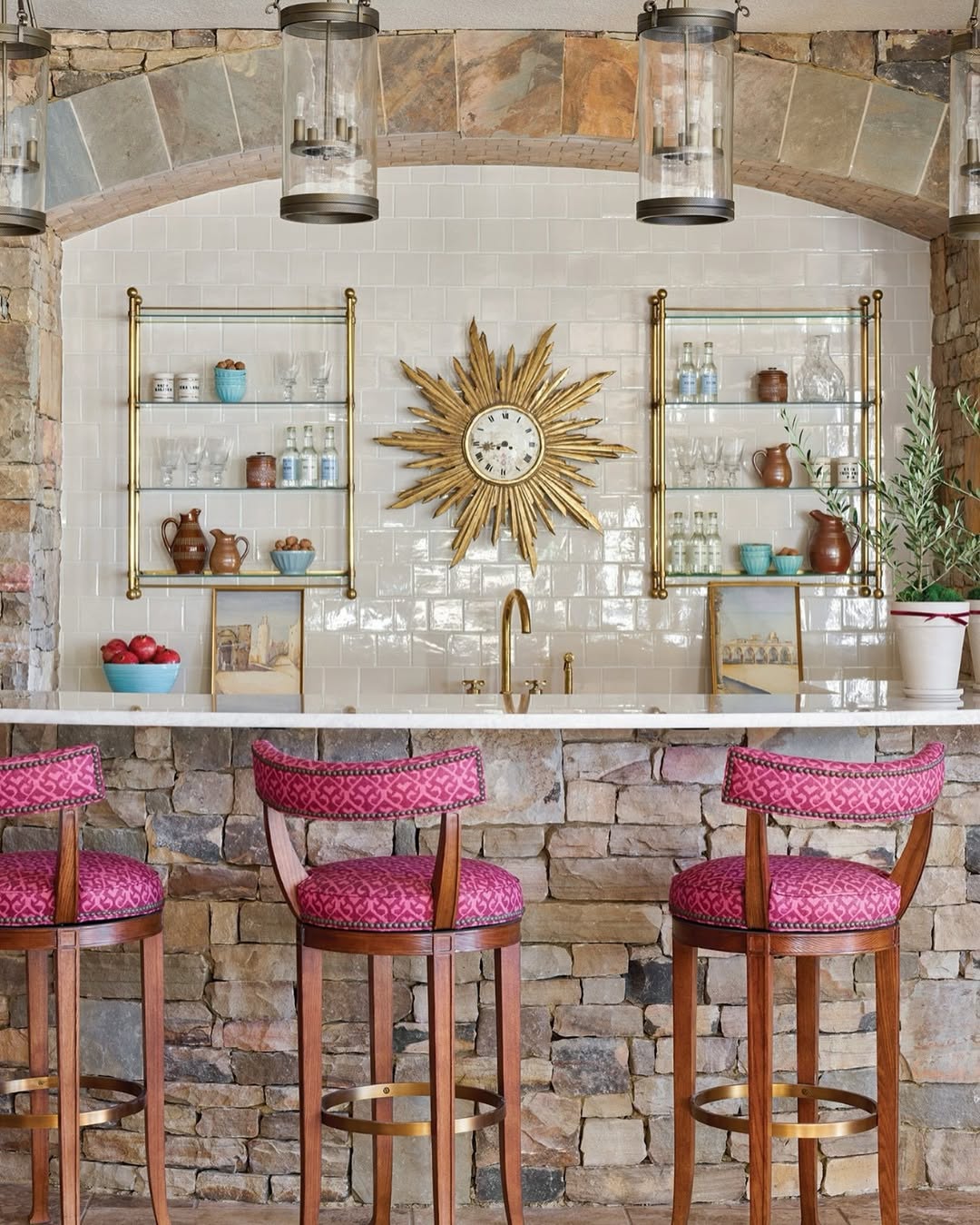 A stone home bar with three pink upholstered stools, a white countertop, a gold sunburst clock, open shelves with dishes, and pendant lights hanging from a stone arch—perfect for stylish home bar ideas.