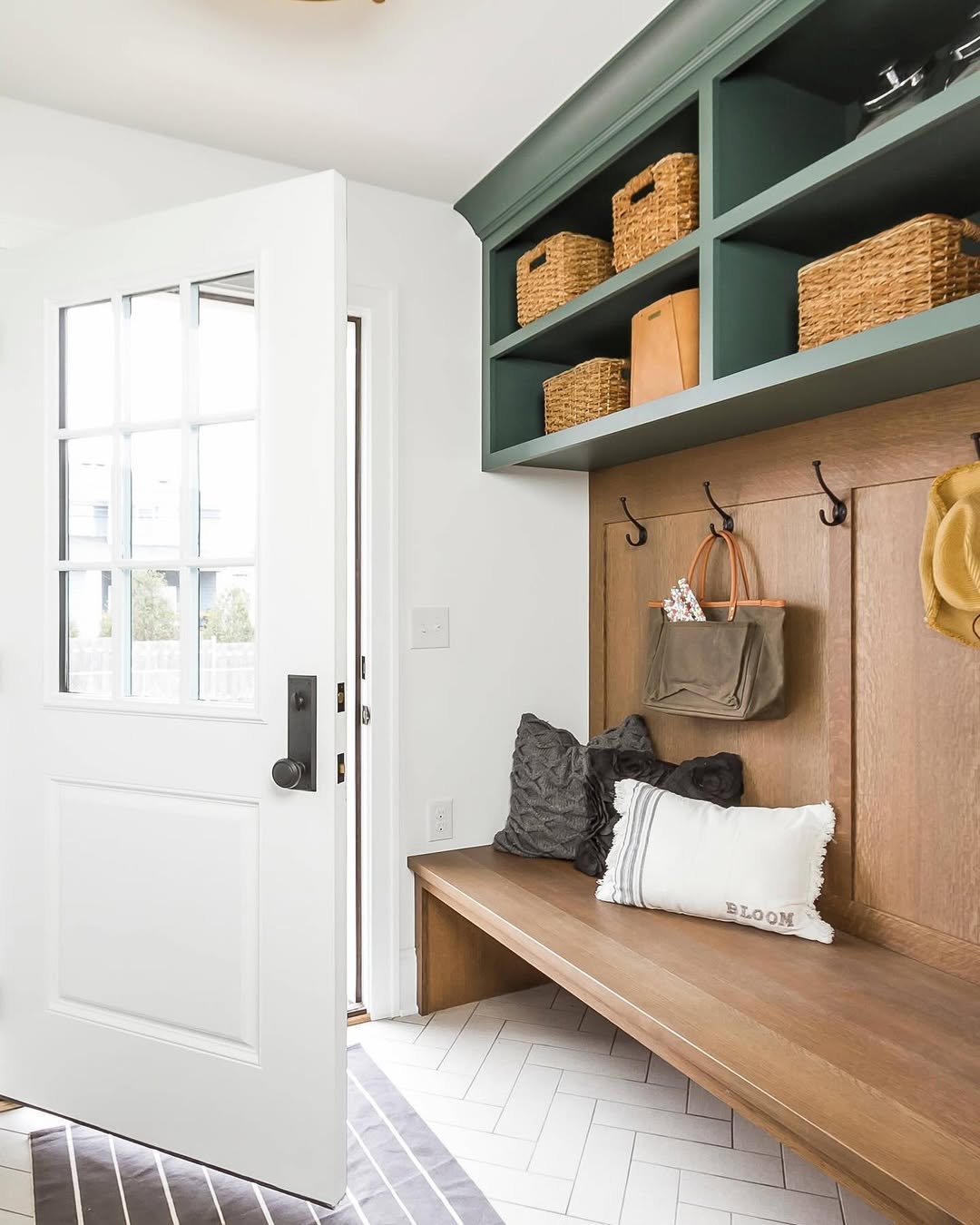 A genius mudroom with a wooden bench, wall hooks holding bags and a hat, woven baskets on green shelves, and a white door open to the entryway. Pillows rest on the bench for added comfort.