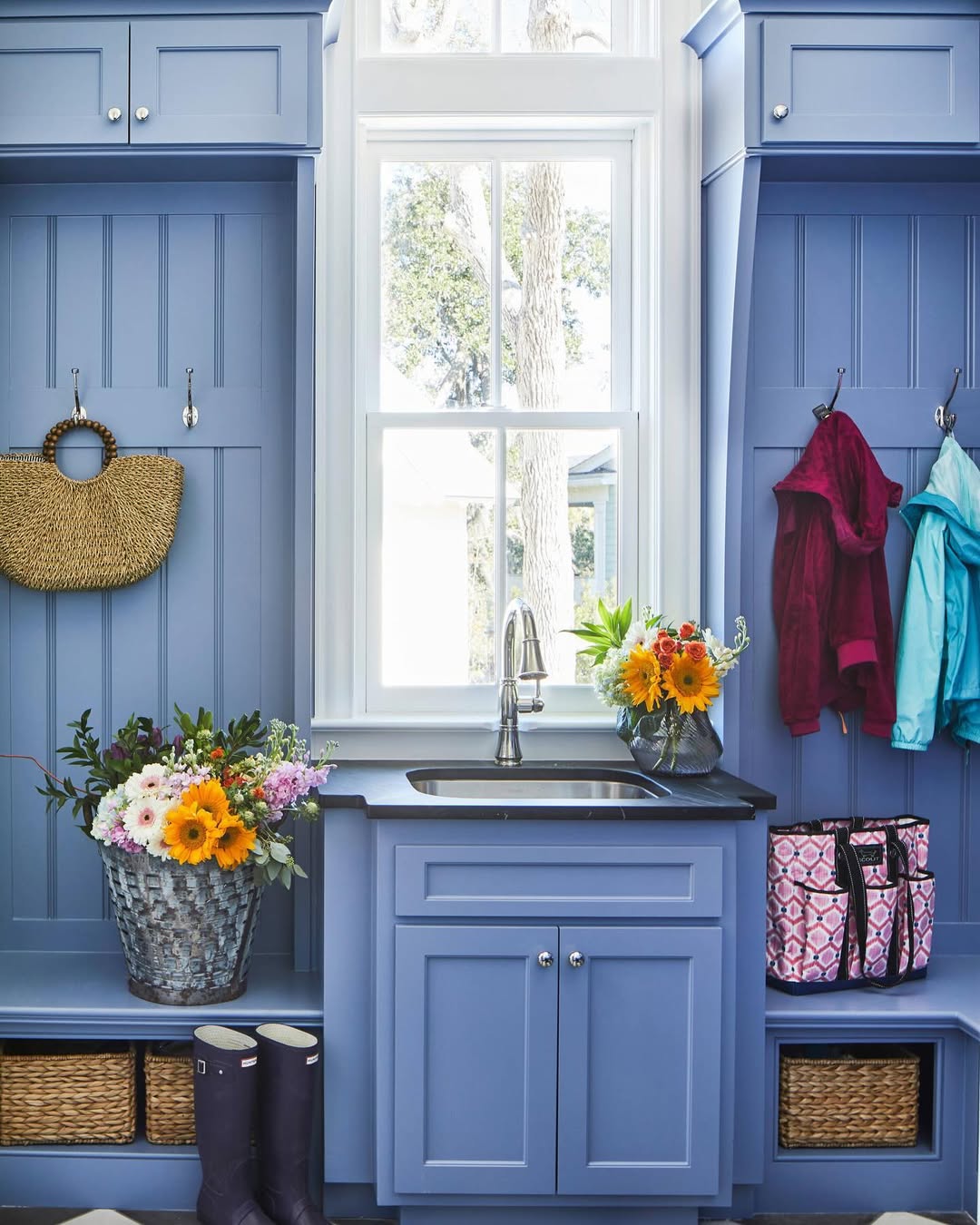 A genius mudroom with blue cabinets, baskets, outerwear, rain boots, fresh flowers in baskets and a vase, a straw bag, and a window above a small sink is the perfect entryway inspiration.