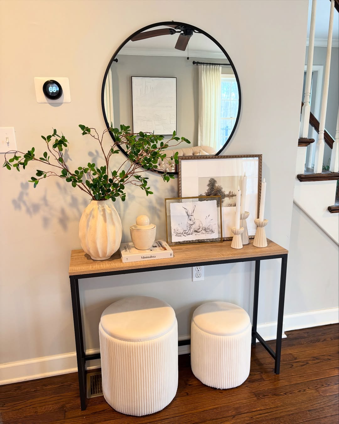 A console table with a vase of greenery, framed art, candles, and stacked books sits below a round mirror, with two cream ottomans underneath—showcasing effortless console table decor for a modern touch.