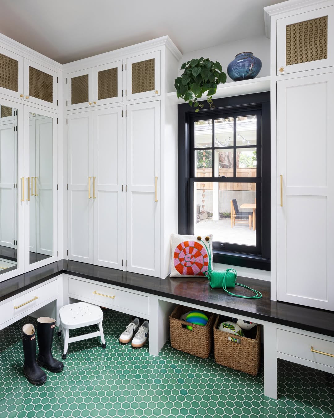 A genius white entryway mudroom with black countertops, green hex tile floor, storage cabinets, baskets, shoes, boots, a step stool, a plant, and a window looking out to the patio.