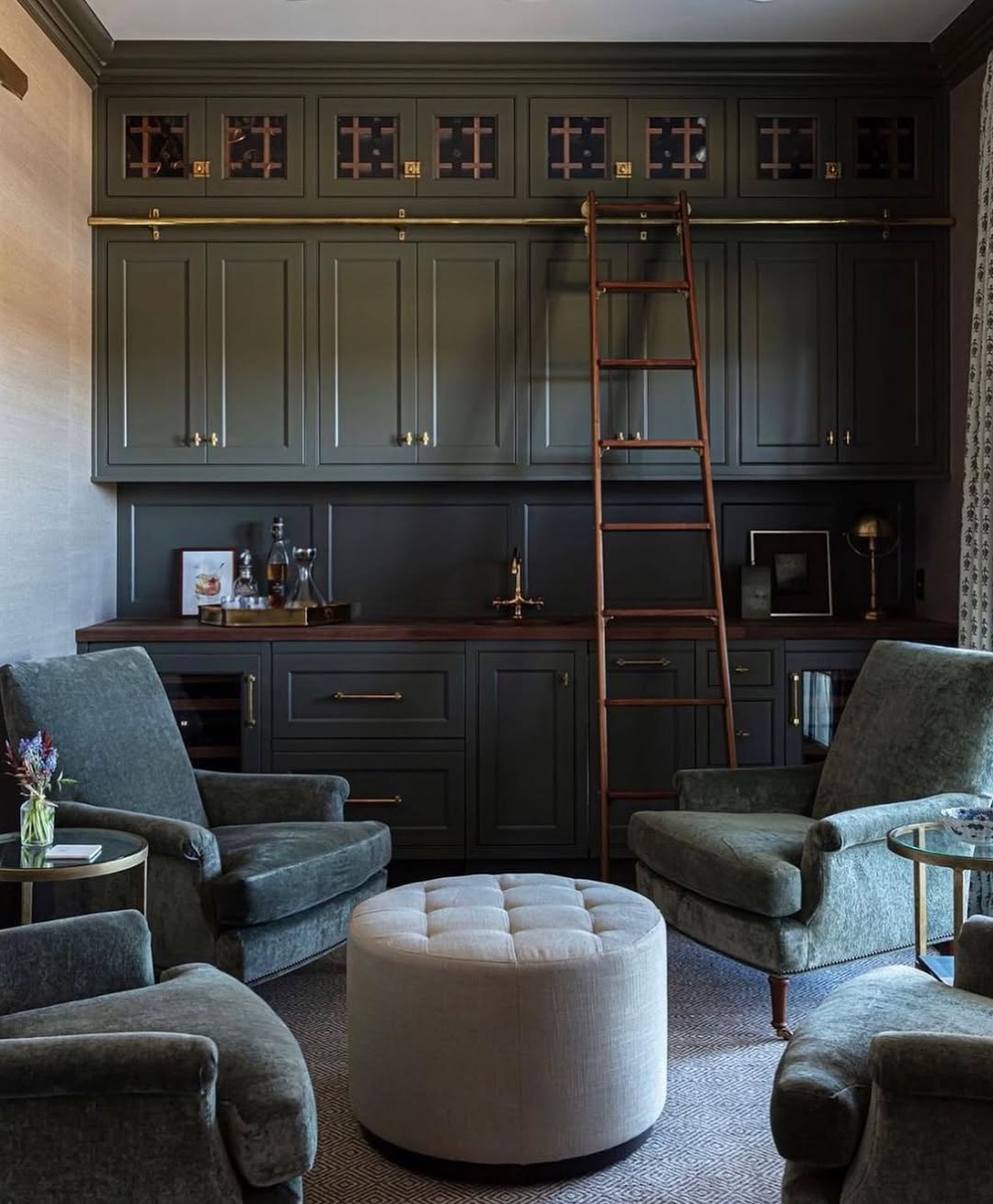 A home library or lounge with green cabinets, a wooden countertop, a rolling ladder, two green armchairs, and a round tufted ottoman—perfect for relaxing or exploring gorgeous bar ideas for home. A side table with flowers completes the inviting space.