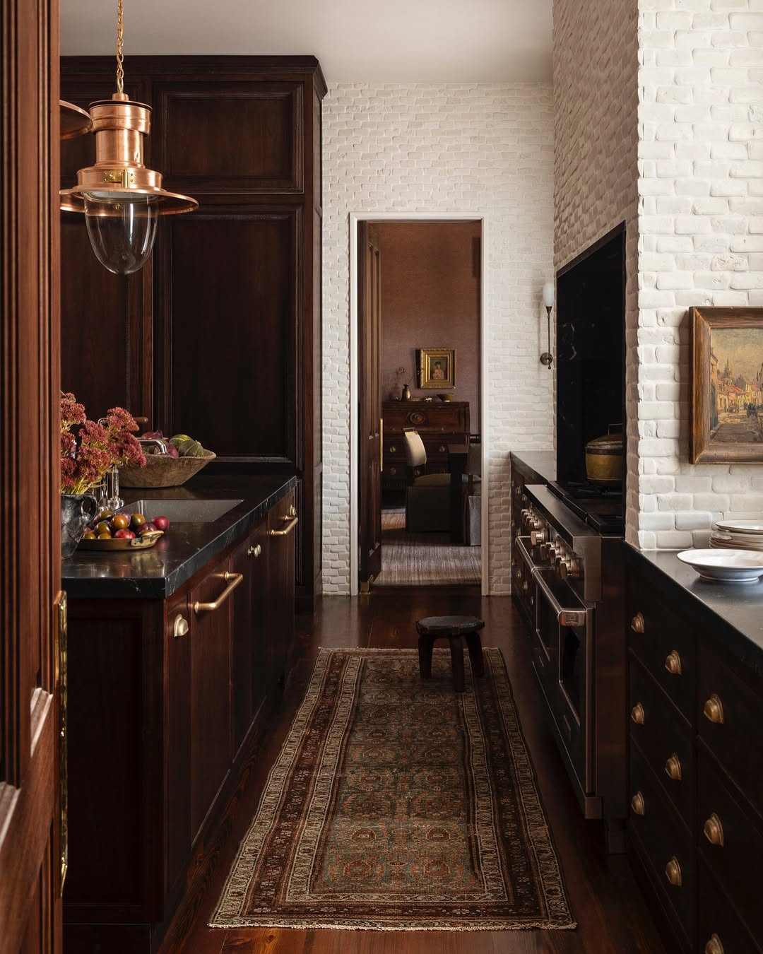 A kitchen with dark wood cabinets, brass hardware, a patterned rug, fruit on the counter, and white brick walls, leading to another room through an open doorway.