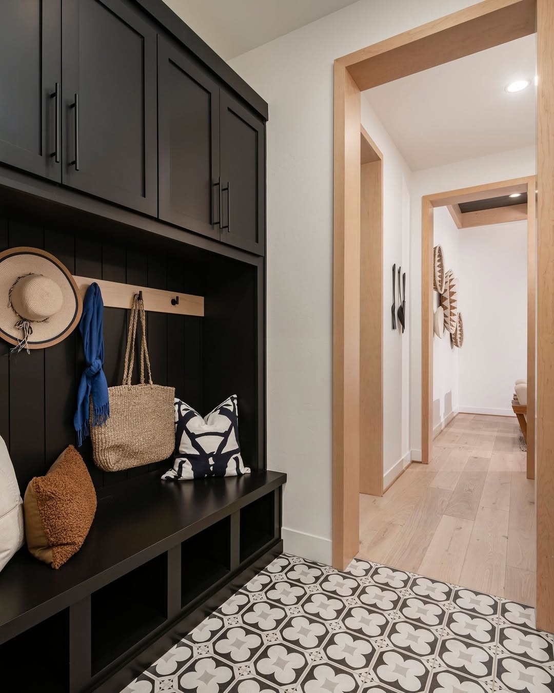 A hallway with a patterned tile floor, black built-in bench and cabinets, hooks with hats and bags, and light wood door frames offers genius mudroom ideas for a functional entryway.